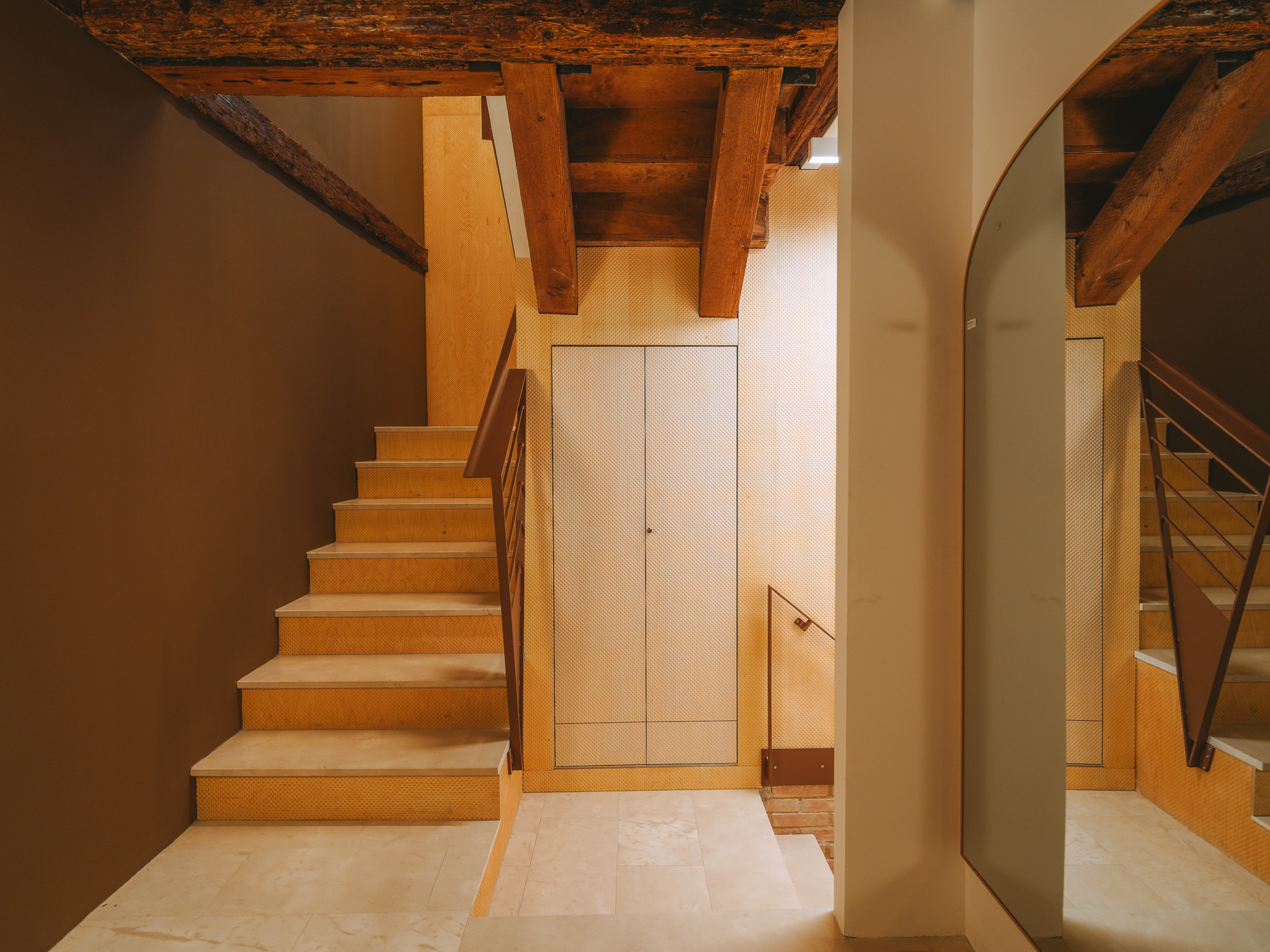 Pale stone staircase with a dark metal handrail, exposed wooden beams and an arched mirror.